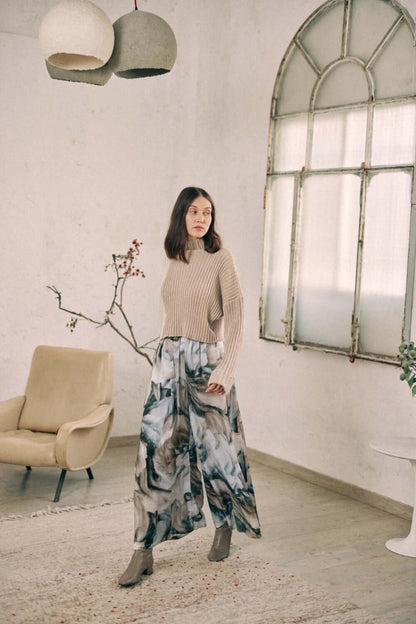 Woman wearing printed wide trousers made from Ecovero viscose, styled with a beige sweater, standing in a stylish, minimalist room.