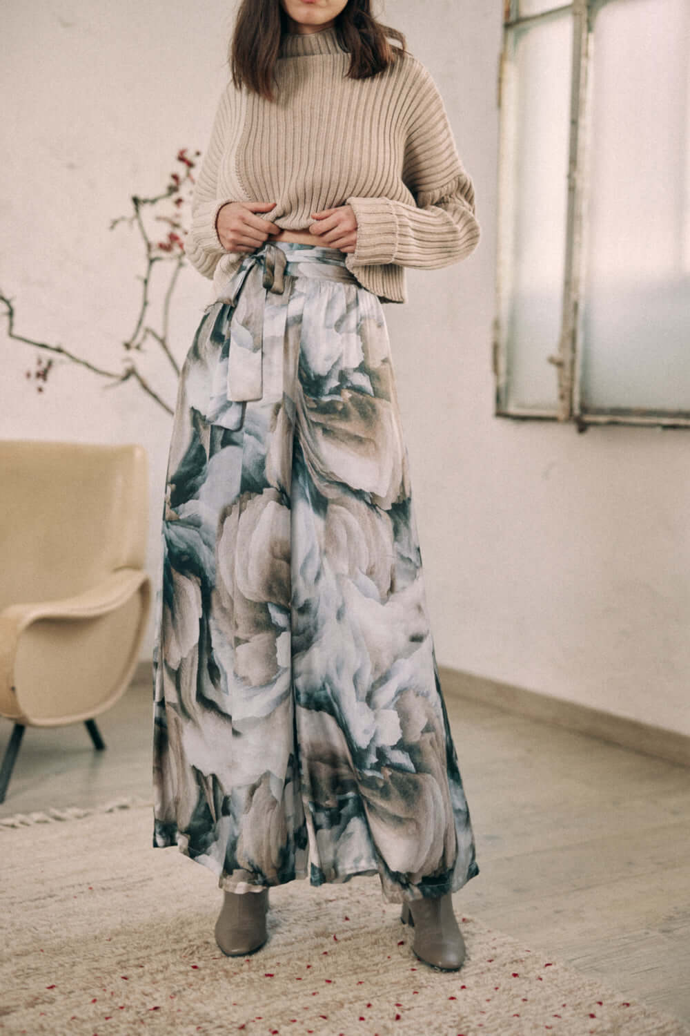 Woman wearing printed wide trousers in Ecovero viscose, featuring autumn floral design and separate belt, styled with a beige sweater.