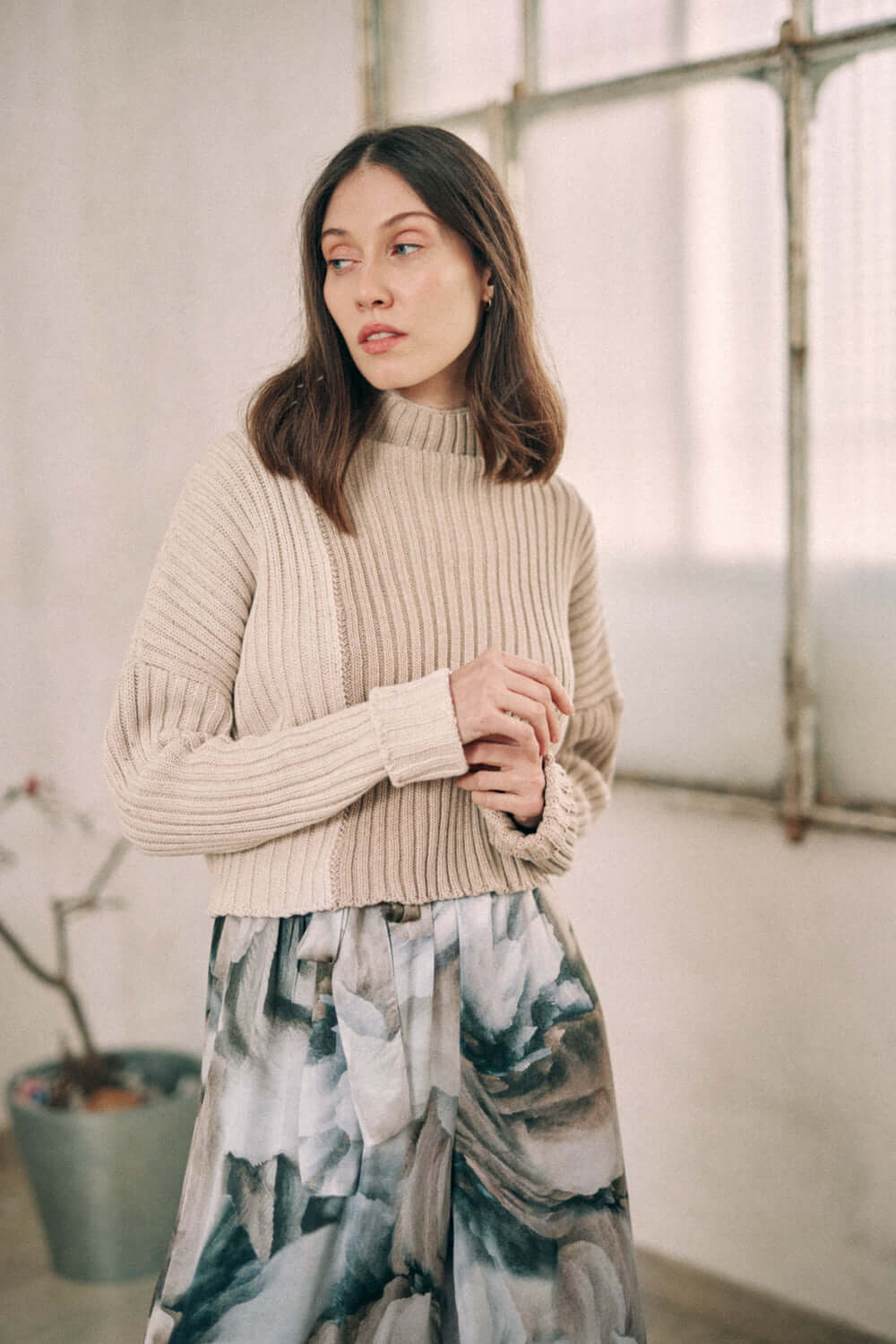 Woman wearing a bicolor beige sweater made from organic cotton, featuring a high neck and cropped wide fit, standing indoors.