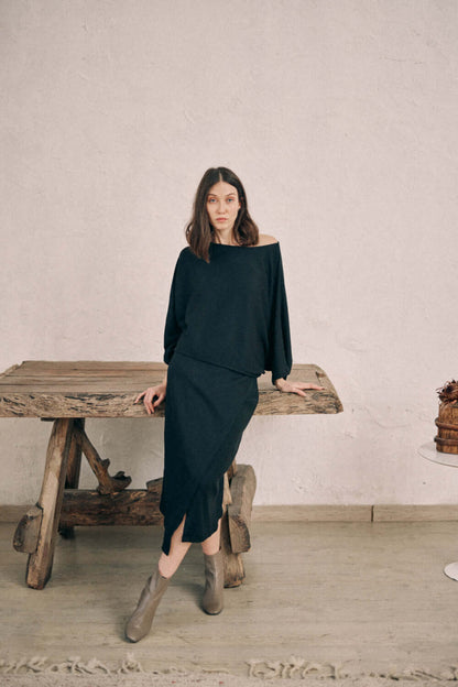 Model wearing black batwing top and matching wrap skirt crafted in thick jersey fabric, leaning against a rustic wooden table.