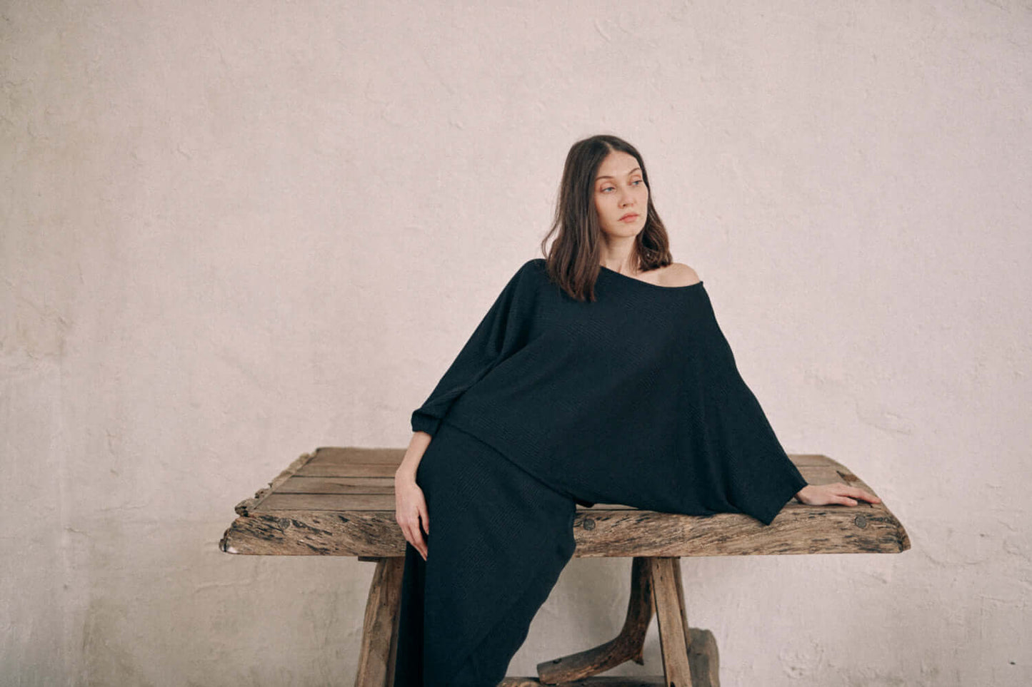 Woman wearing black batwing top crafted from recycled polyester and organic cotton, paired with matching skirt, leaning on wooden table.