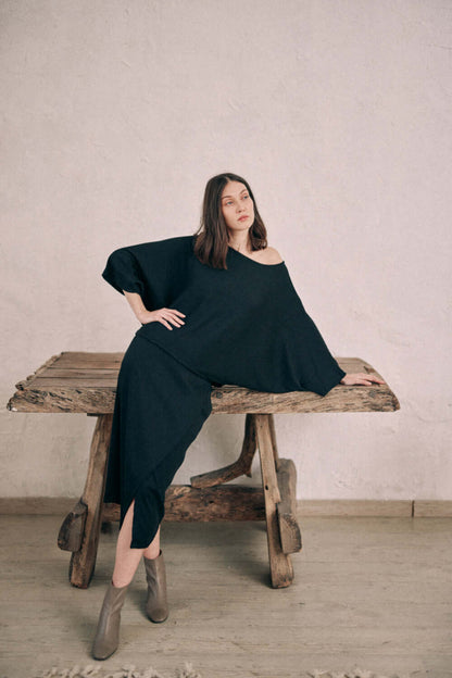 Model wearing black batwing top in thick jersey fabric, paired with matching wrap skirt, standing against rustic table.