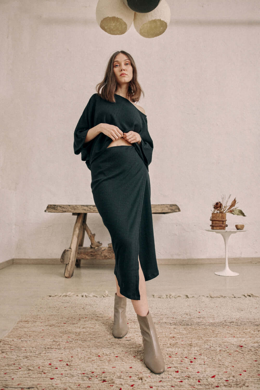 Woman modeling black wrap skirt in thick jersey fabric, paired with matching top and ankle boots, standing in rustic setting.