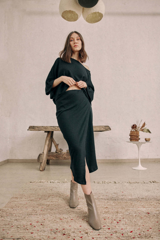 Woman modeling black wrap skirt in thick jersey fabric, paired with matching top and ankle boots, standing in rustic setting.