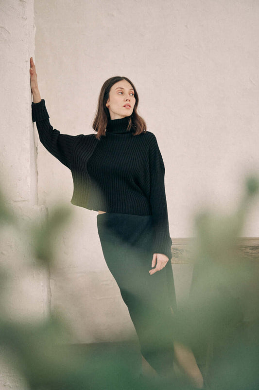 Woman wearing a black bicolor sweater made from organic cotton yarn, featuring a high neck and wide fit, standing against a white wall.