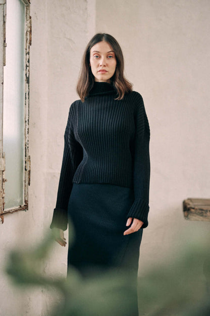 Woman wearing bicolor black sweater made from organic cotton yarn, showcasing a high neck and wide fit design, standing indoors.