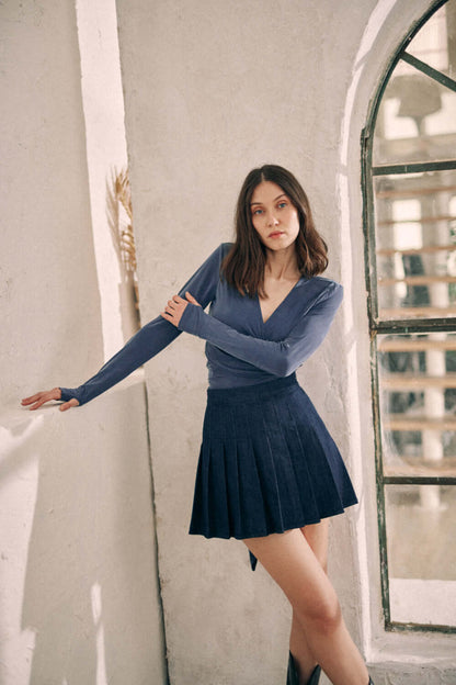 Woman wearing navy cupro wrap top with thumb holes, paired with pleated skirt, standing near arched window in an airy room.