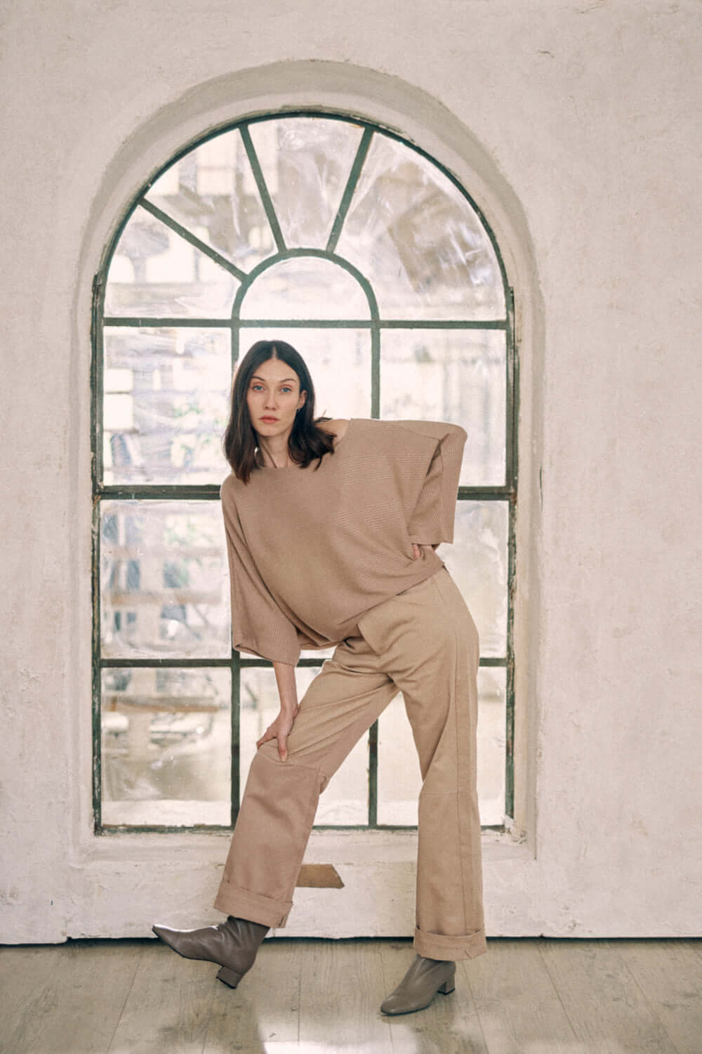 Woman wearing taupe batwing top and trousers set, standing in front of arched window, showcasing wide body design and thick jersey fabric.