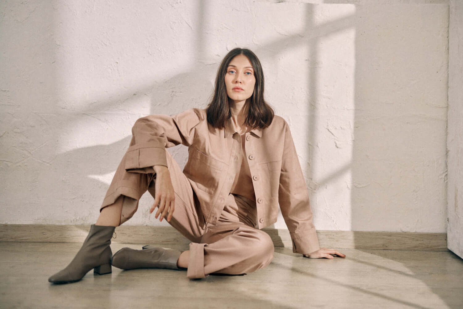 Model wearing taupe denim jacket and matching pants, sitting on floor in a minimalist setting, showcasing organic cotton fabric and COSSAC buttons.